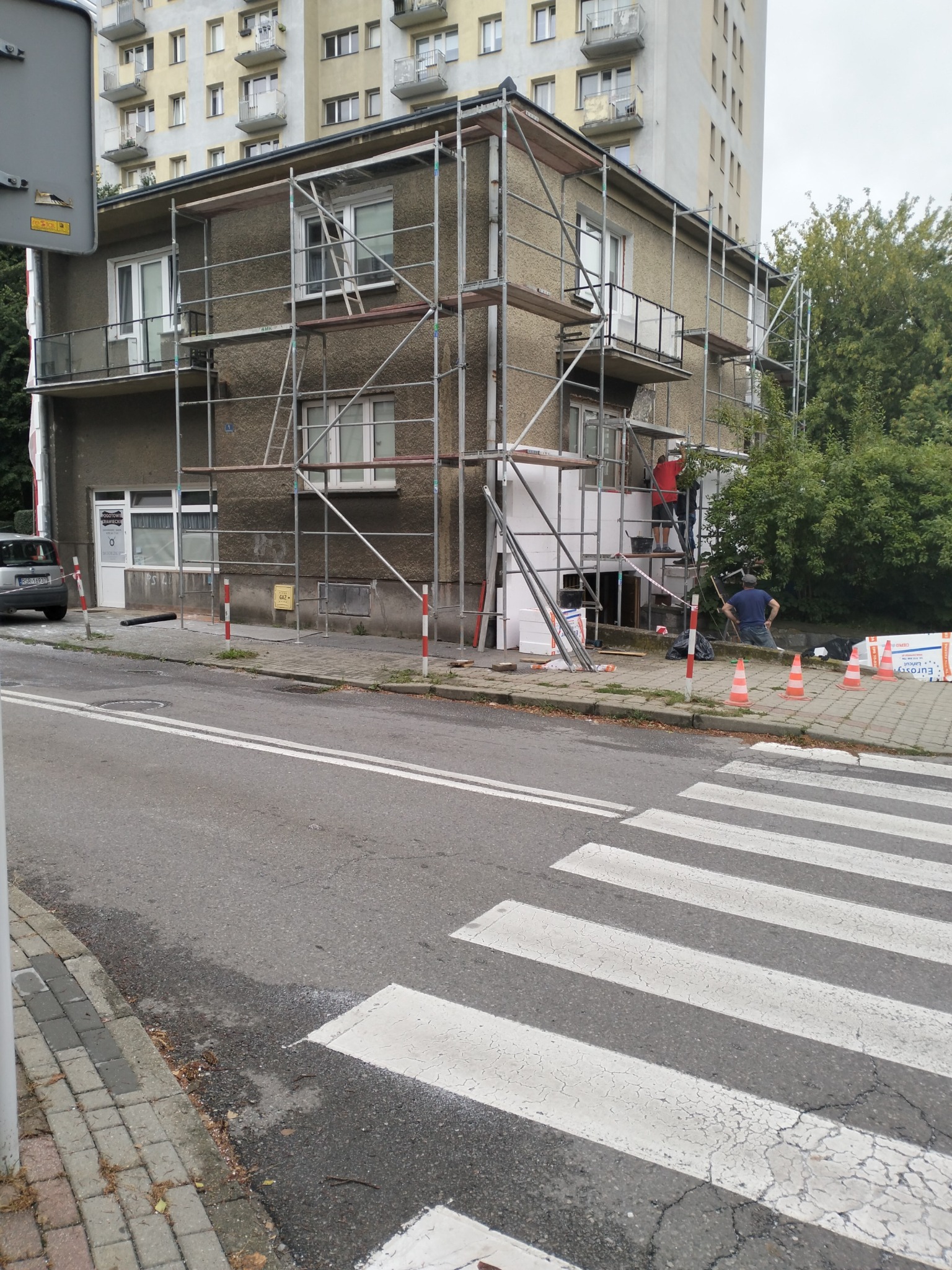 Dwupiętrowy budynek w trakcie prac elewacyjnych, z widocznym rusztowaniem, pracownikami na rusztowaniu oraz stosami białego styropianu na chodniku, na tle bloku mieszkalnego i przejścia dla pieszych.