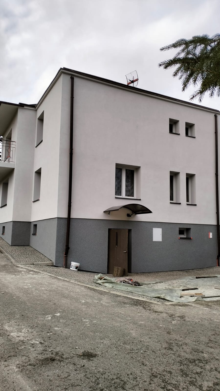 Dwukolorowa elewacja budynku z szarą dolną częścią i białą górną, widoczne okna, drzwi wejściowe z daszkiem, rynna oraz antena na dachu.