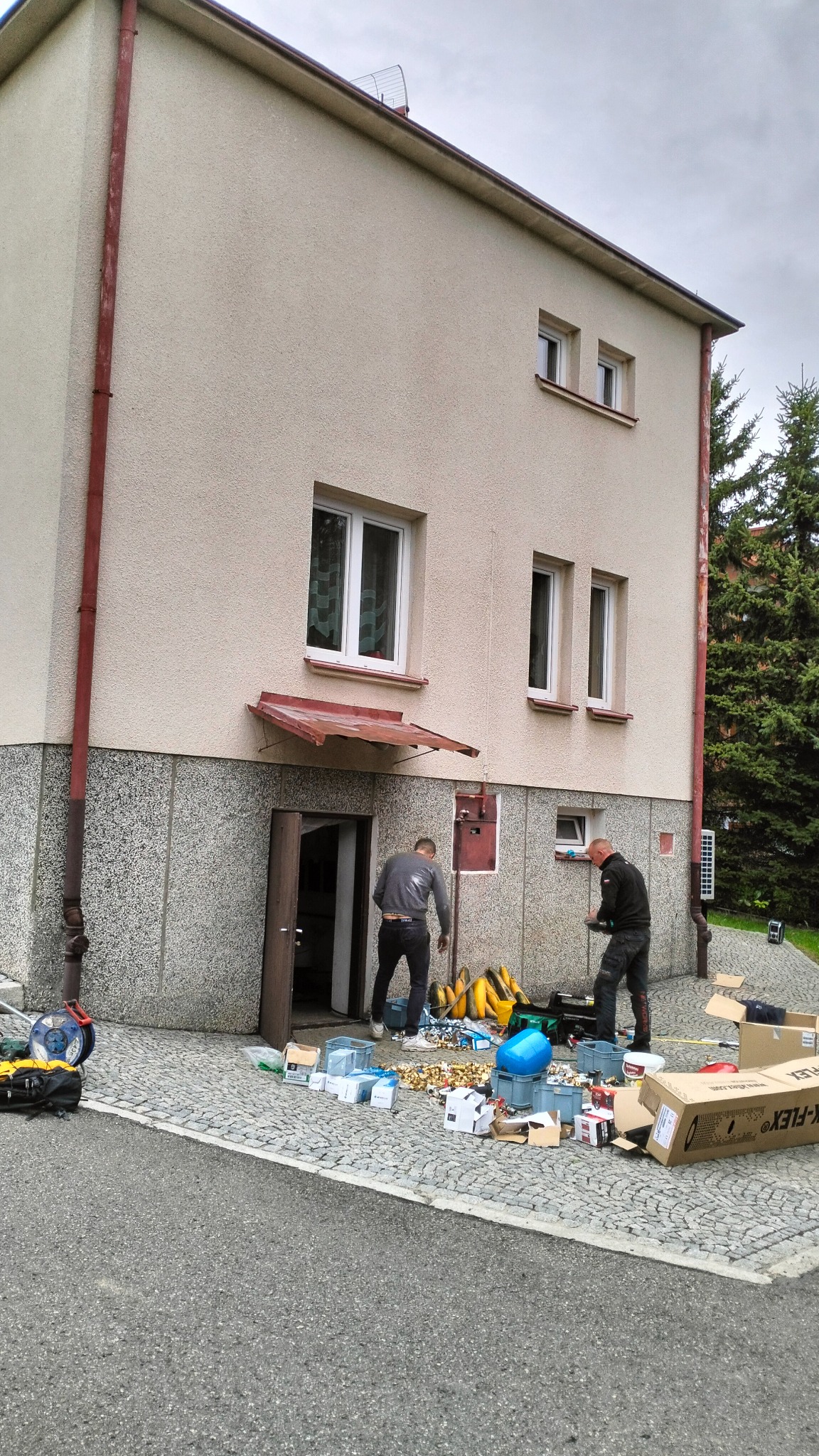 Dwóch mężczyzn przy budynku z elewacją, rozpakowujących materiały hydrauliczne na chodniku z kostki brukowej, widoczne skrzynki, rury i narzędzia.