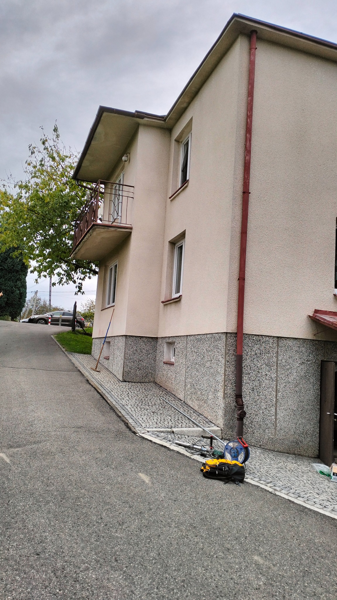 Dwukondygnacyjny dom z beżową elewacją i szarą kamienną podmurówką, widoczny z poziomu ulicy, z narzędziami i sprzętem leżącym na chodniku przed budynkiem. Czerwona rynna biegnie wzdłuż ściany.