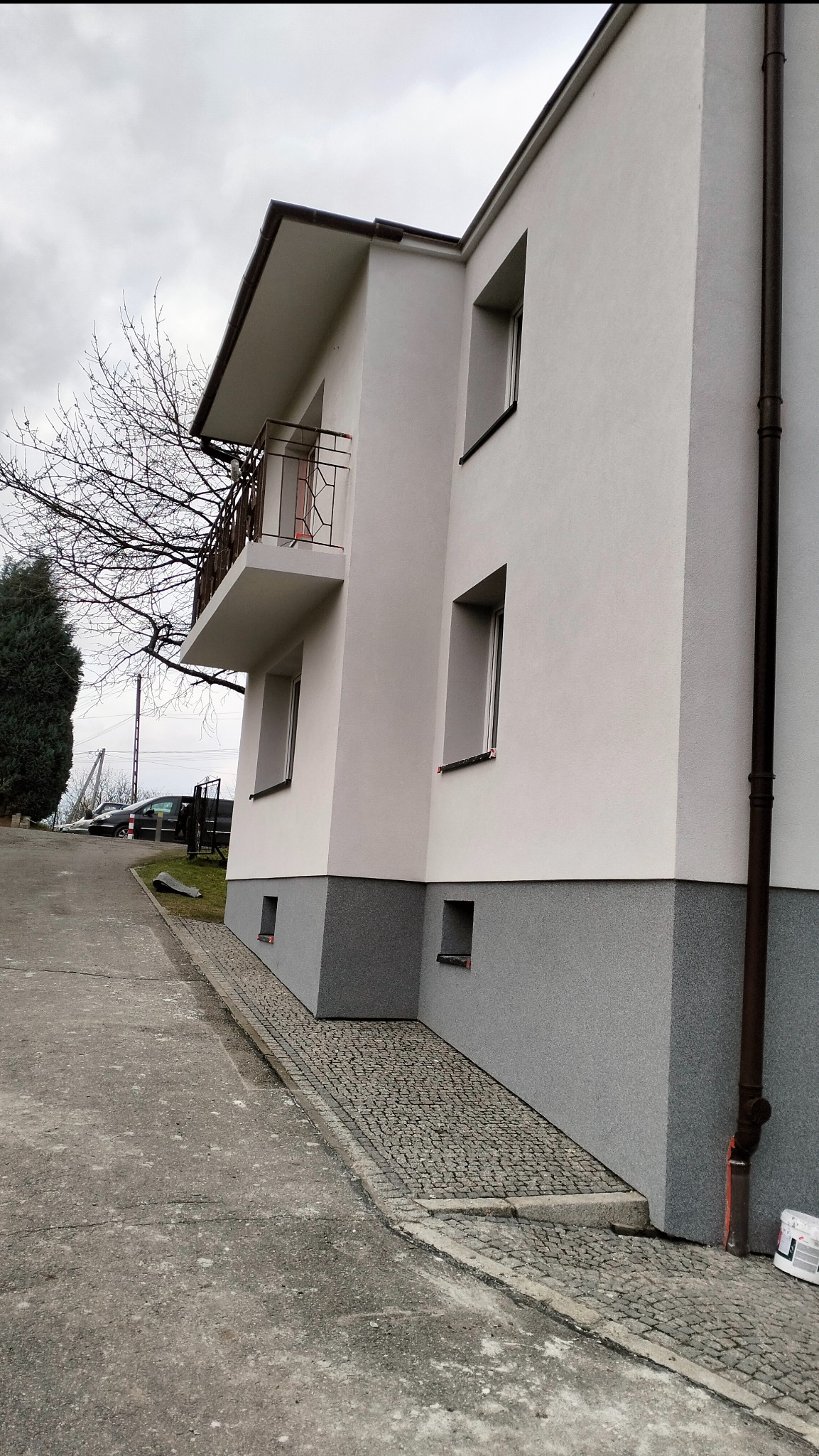 Dwukondygnacyjny budynek z balkonem po remoncie elewacji, z szarą, ziarnistą fakturą w dolnej części i białą powyżej, brązowe rynny i obramowania okien, widok z dołu na pochmurne niebo.