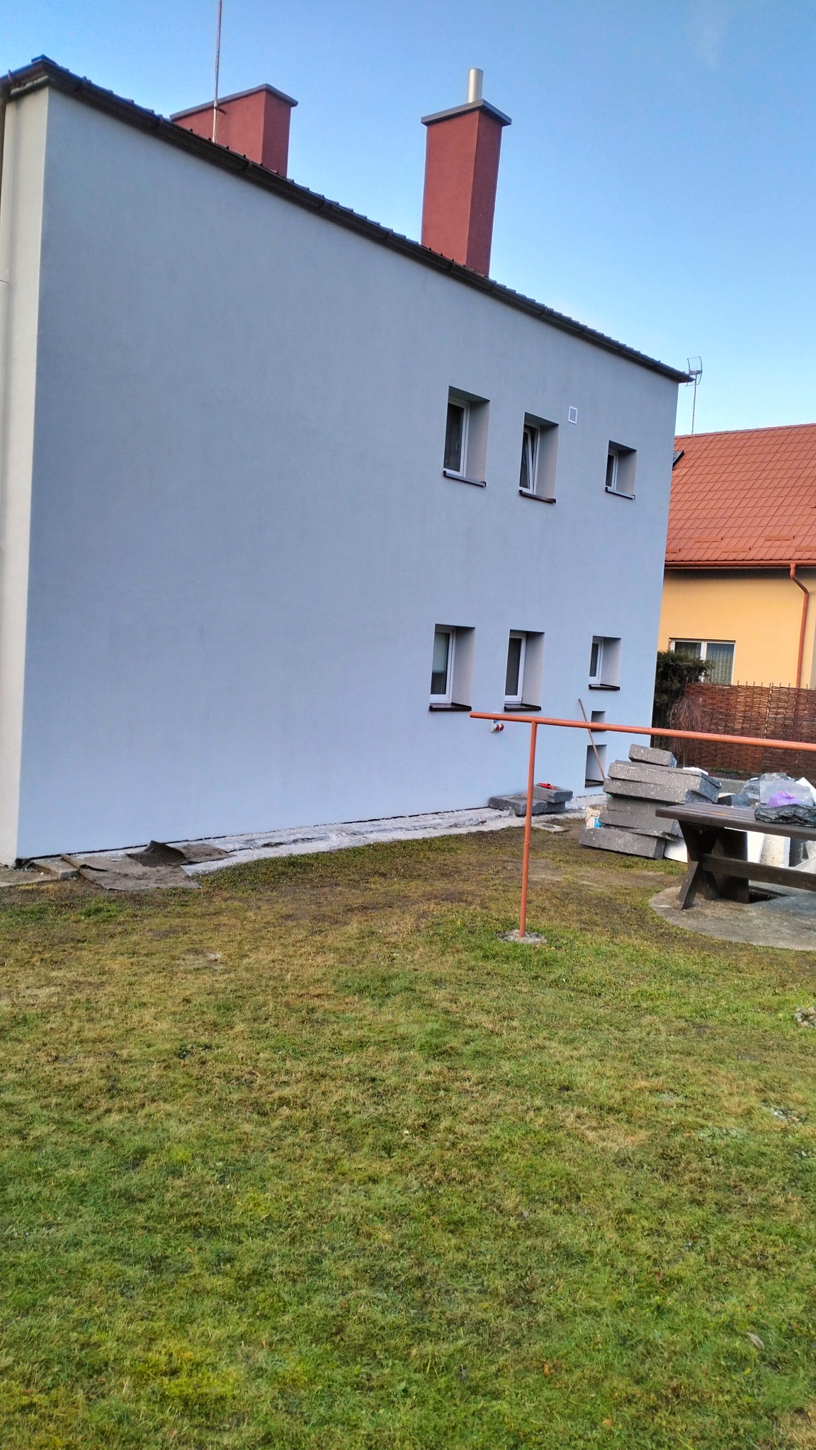 Świeżo pomalowana ściana budynku z widocznymi oknami, czerwonymi kominami i fragmentem trawnika z elementami budowlanymi w tle.