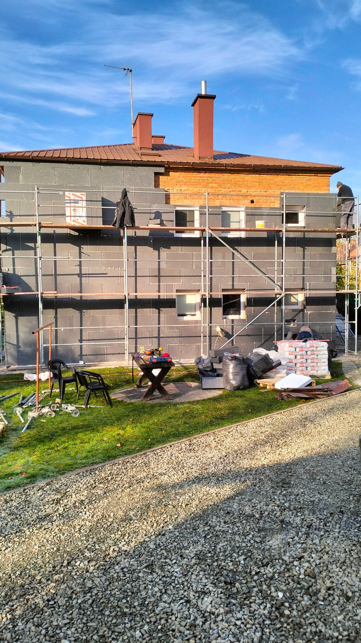 Dom w trakcie ocieplania styropianem, widoczne rusztowania, przygotowane materiały budowlane, narzędzia na stole i pracownicy na rusztowaniu, częściowo ocieplona ściana budynku.