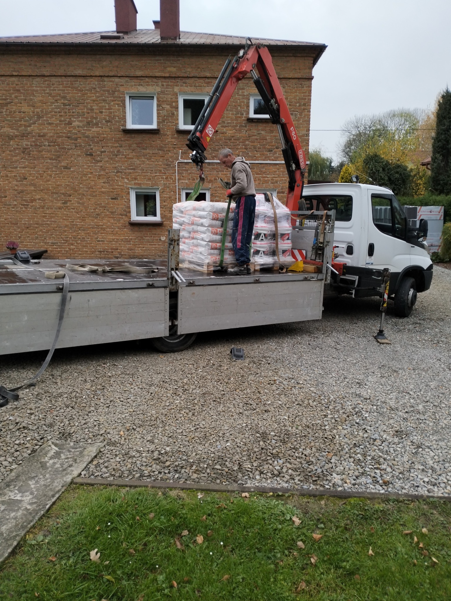 Rozładunek worków z zaprawą elewacyjną za pomocą dźwigu samochodowego na tle ceglanego budynku.