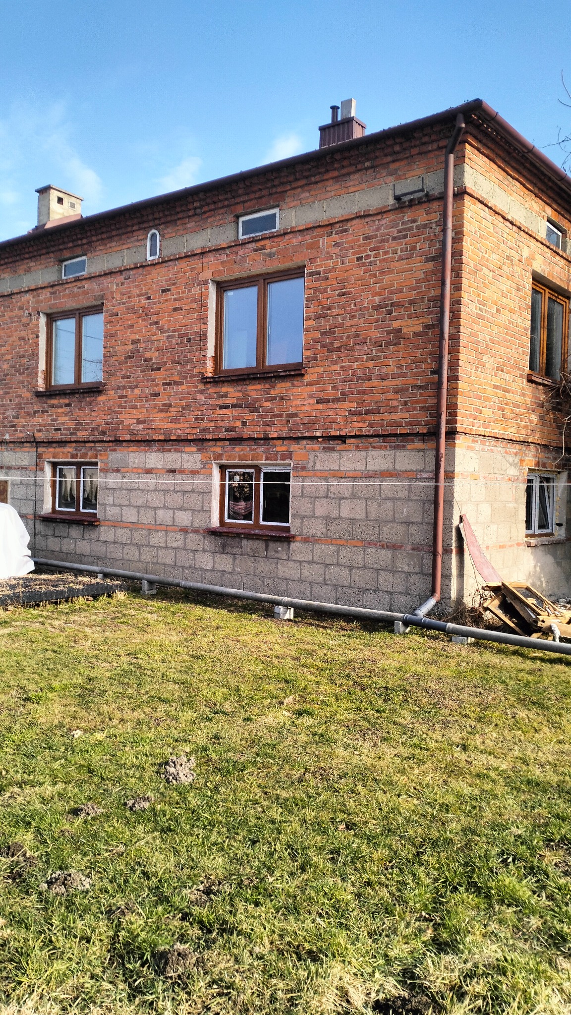 Dwupiętrowy budynek z czerwonej cegły z szarą, betonową podstawą, widoczne okna z drewnianymi ramami, rury spustowe i trawnik przed budynkiem.