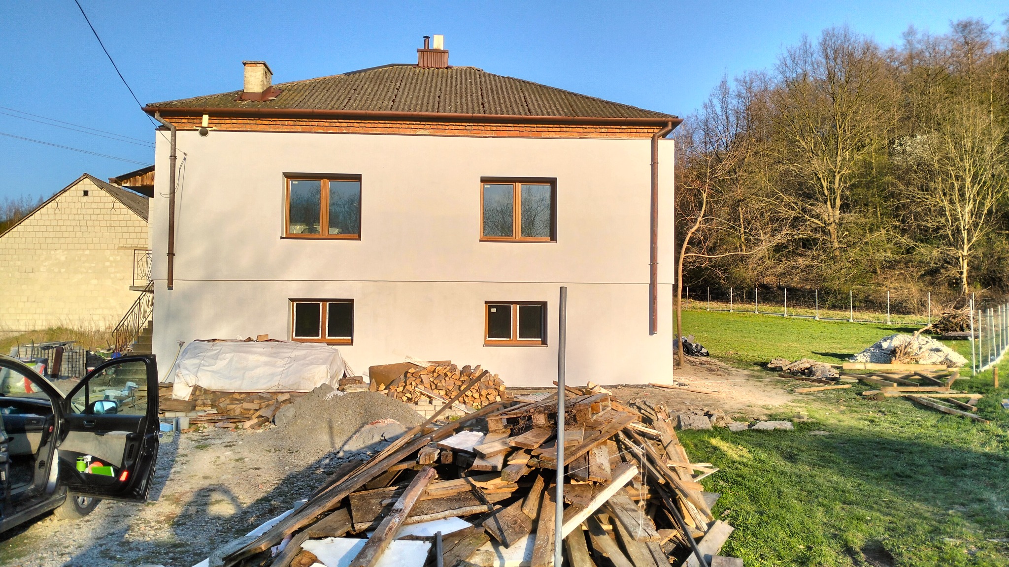 Dwupiętrowy dom z oknami z brązowymi ramami po remoncie elewacji w Rzeszowie, z widocznym stosem drewna i otwartymi drzwiami samochodu na pierwszym planie.