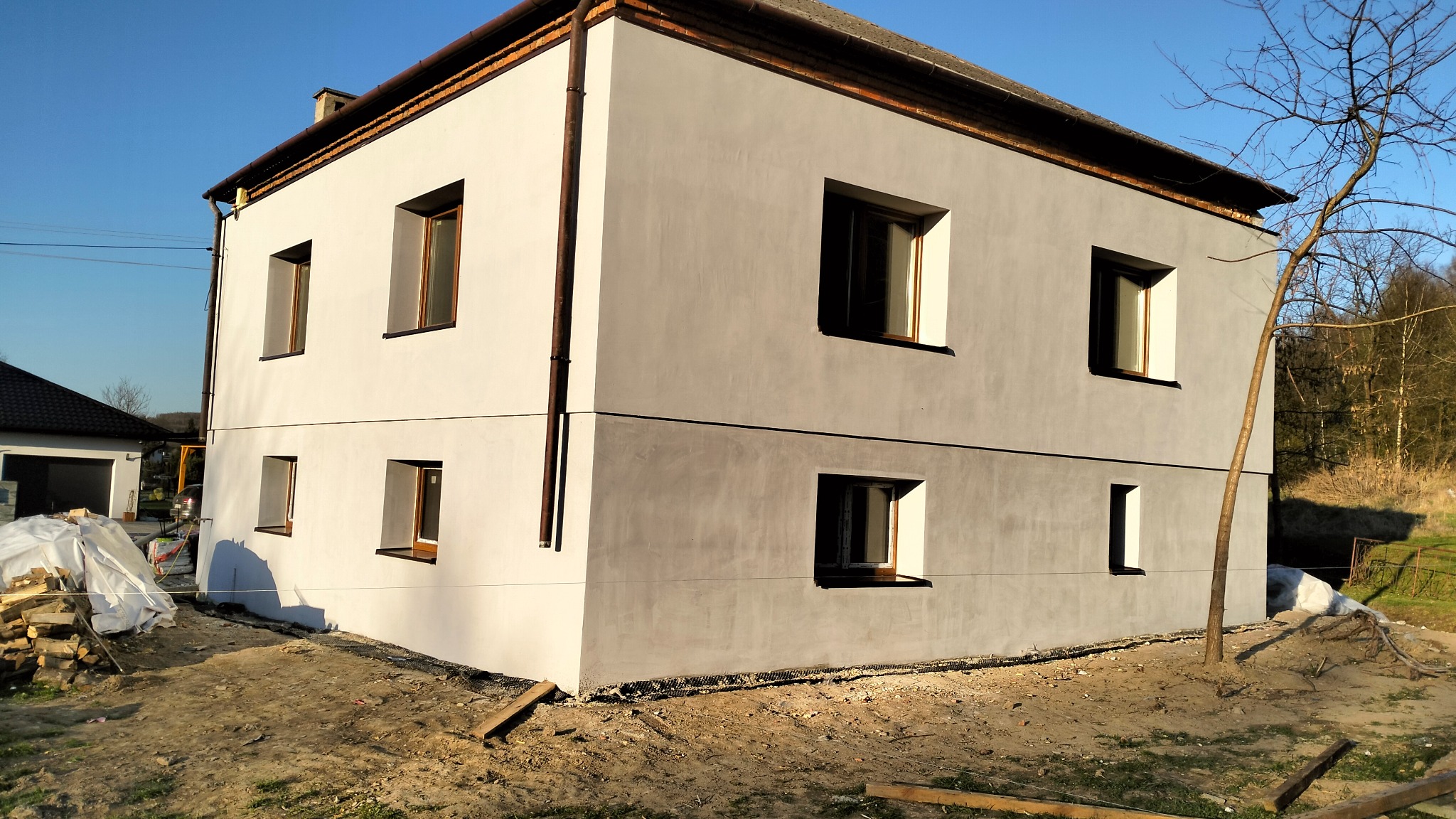 Dwukondygnacyjny dom z szarą elewacją, widoczne brązowe okna i orynnowanie, teren wokół domu w trakcie prac wykończeniowych.