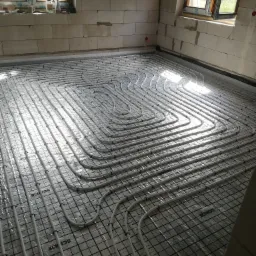 Układanie rur ogrzewania podłogowego na folii z nadrukiem producenta, widoczne okna i surowe ściany z betonu kom&oacute;rkowego.