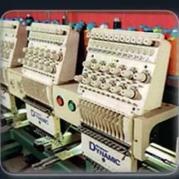 Rząd automatycznych maszyn do szycia z wieloma igłami, z widocznym systemem nawlekanych nici w różnych kolorach, ustawionych w rzędzie w warsztacie.