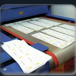 Białe etykiety z nadrukiem na taśmie produkcyjnej, gotowe do odcięcia, z widocznym fragmentem maszyny.