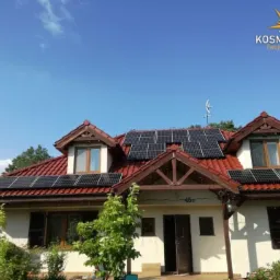Dom jednorodzinny z czerwoną dachówką, na której zamontowano panele słoneczne; widoczny także mały wiatrak na dachu i logo firmy instalującej.