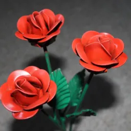 Trzy czerwone, metalowe róże z zielonymi łodygami i liśćmi na rozmytym, szarym tle. Kwiaty wykonane z blachy, widoczne ślady spawania.