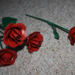 Czerwone, metalowe róże spawane, z zielonymi łodygami, leżące na dywanie o neutralnym kolorze.