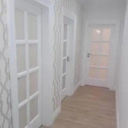 Korytarz z trzema białymi skrzydłami z przeszkleniami, drewniana podłoga i tapeta w geometryczny wzór.