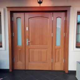 Drewniane wejściowe z bocznymi przeszkleniami, wkomponowane w elewację z oknami o podobnej stylistyce, uzupełnione dekoracyjnymi roślinami w donicach.