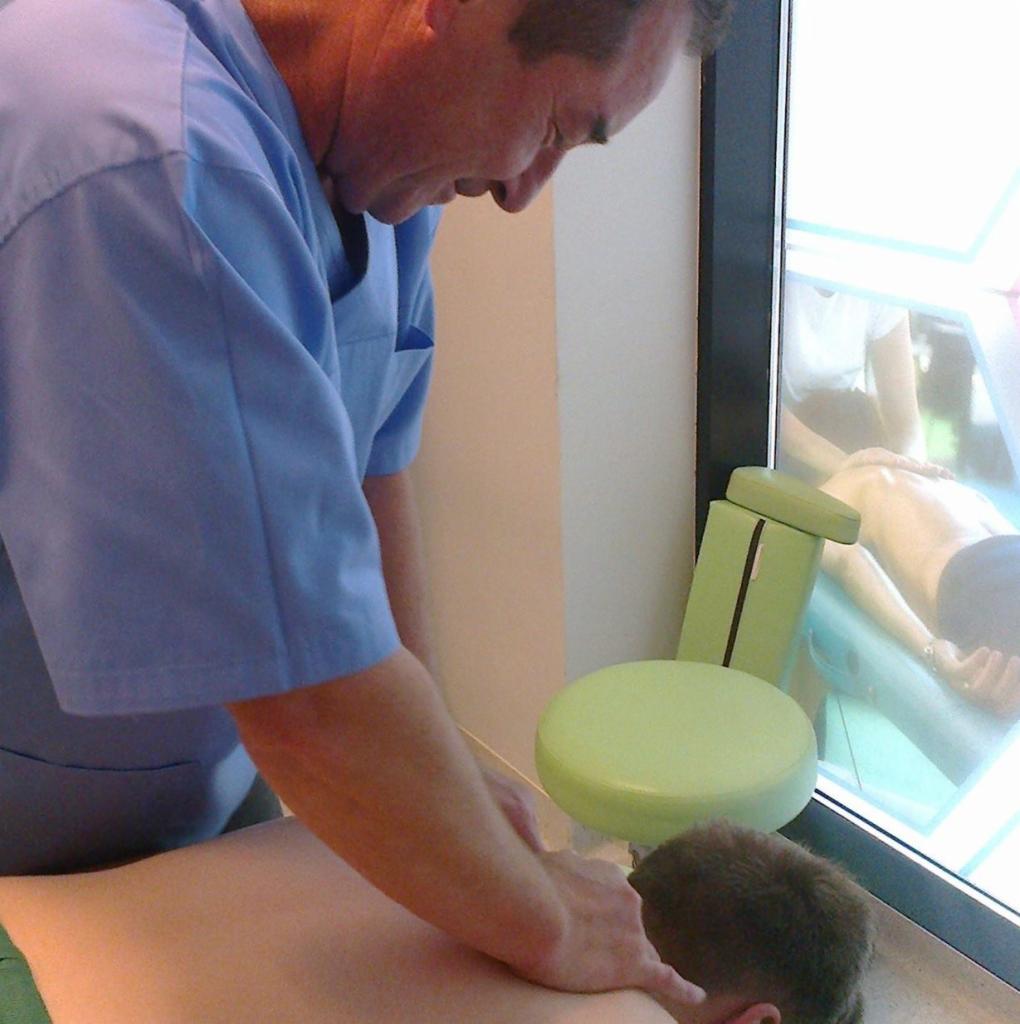 Masaż pleców wykonywany przez mężczyznę w niebieskim uniformie, w tle zielone krzesło i ekran z prezentacją innej sesji masażu.