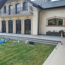 Dom z kutymi elementami balkonów i okien po pracach remontowych, na trawniku widoczne narzędzia i pozostałości materiałów budowlanych.