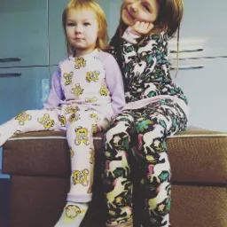 Dwie dziewczynki siedzą na pufie w pasujących piżamach: jedna w jasnofioletowej z motywem misiów, druga w ciemnej z jednorożcami na tle błękitnych szafek.