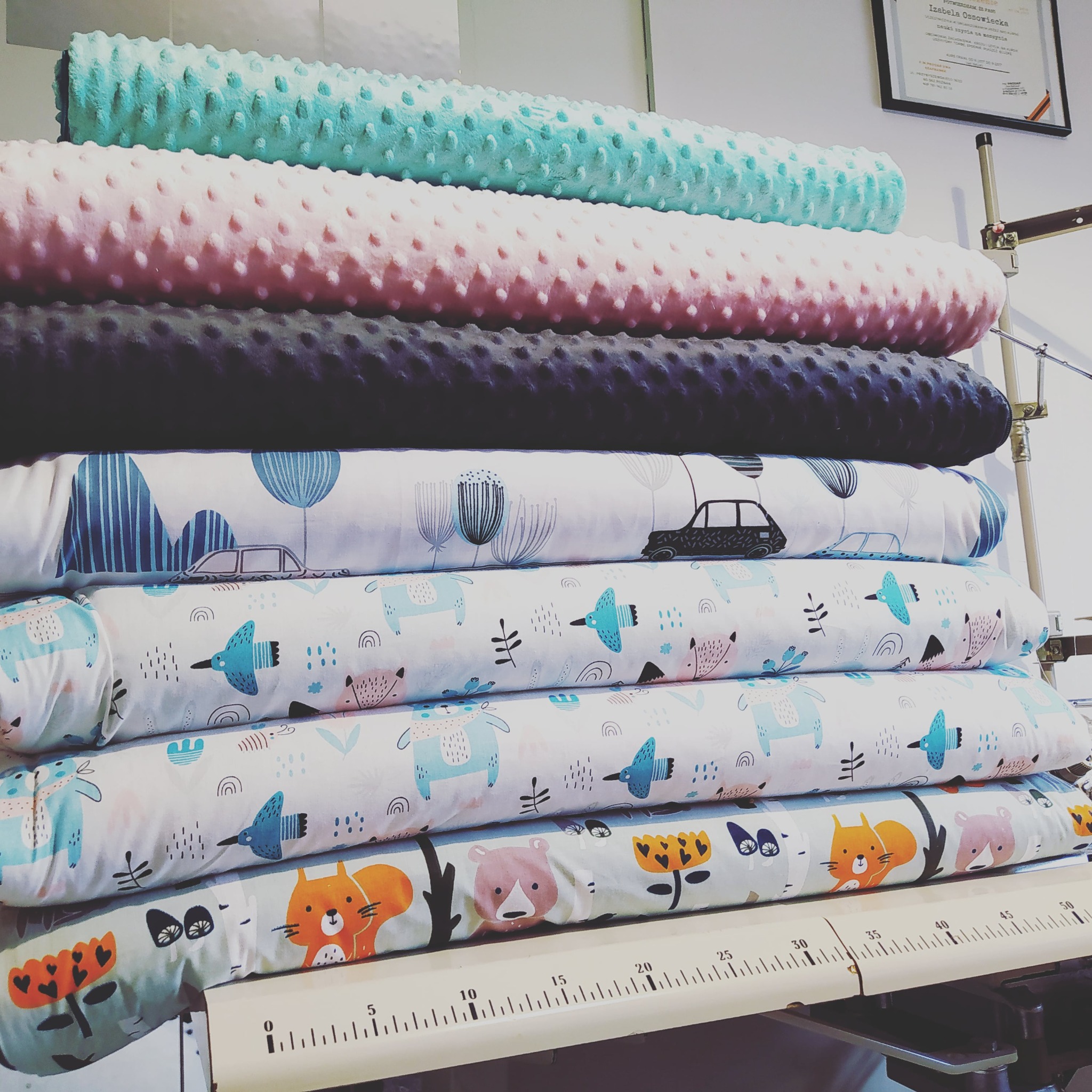 Stos zwiniętych tkanin dziecięcych z wzorami zwierząt i aut, ułożonych na stole z miarką, w tle certyfikat.
