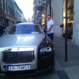 Srebrno-czarny Rolls Royce zaparkowany na brukowanej ulicy w Krakowie, obok stoi mężczyzna w białej koszuli i granatowych szortach.