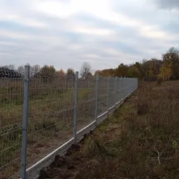 Długi, nowy płot z siatki panelowej na betonowej podmurówce, oddzielający teren z łąką od zalesionego obszaru, widoczny w pochmurny dzień.