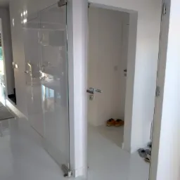 Nowoczesne wnętrze z białymi płytkami podłogowymi, ukazujące szklane drzwi z metalowymi zawiasami oraz białe drzwi z chromowanymi klamkami, z widocznymi butami i pantoflami w tle.