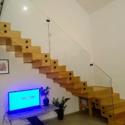 Drewniane schody wspornikowe z balustradą ze szkła mocowanego punktowo, widok z dołu, w tle ściana z telewizorem i dekoracjami.