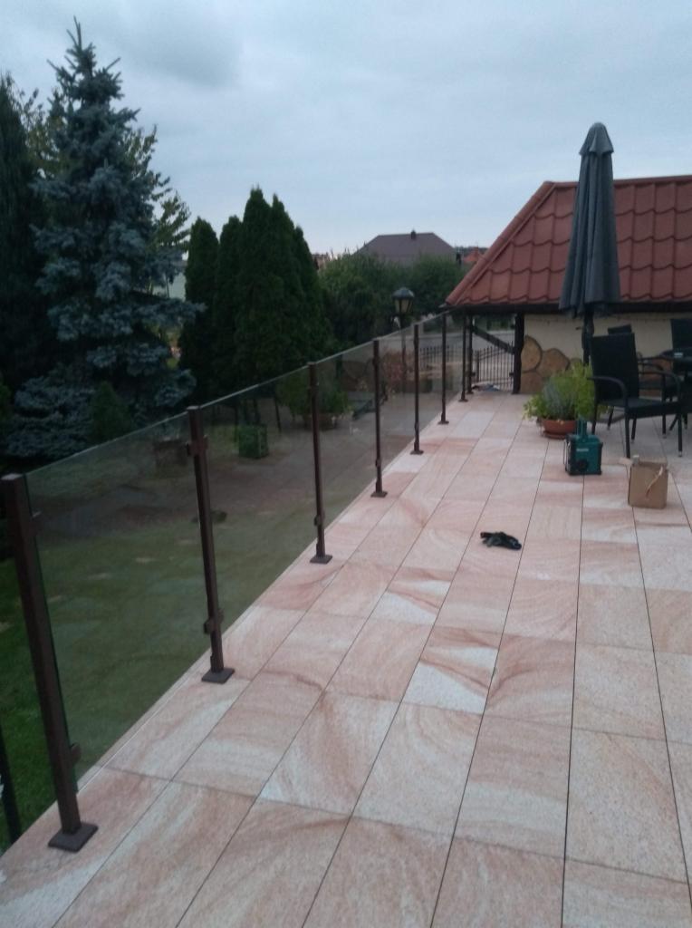 Zewnętrzny taras wyłożony płytkami imitującymi piaskowiec, zabezpieczony szklaną balustradą z brązowymi słupkami, widok na ogród i dom z zadaszeniem.