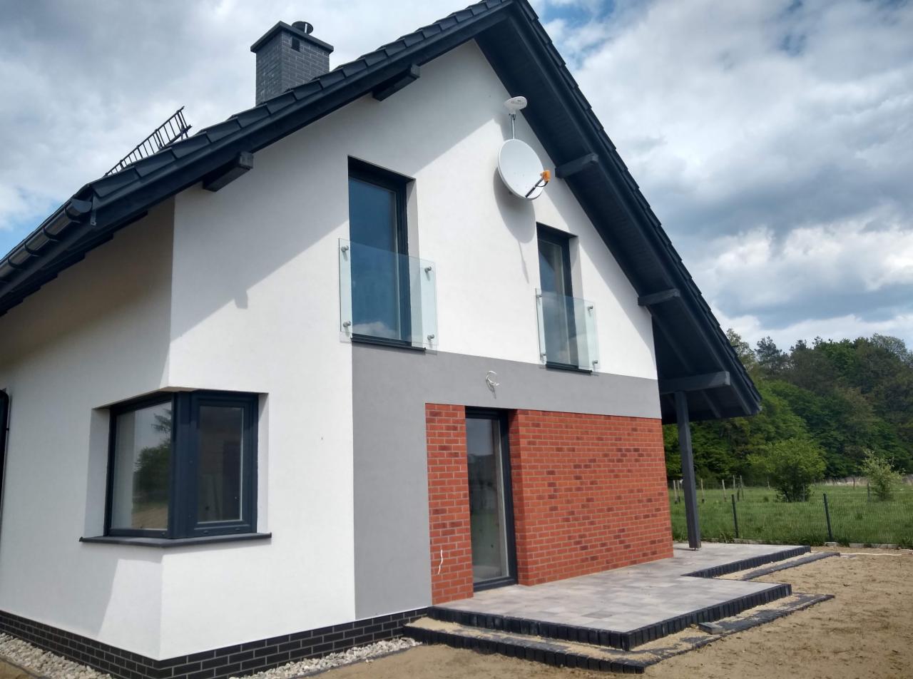 Nowoczesny dom jednorodzinny w Strzyżowie z białymi ścianami, grafitową stolarką okienną i szklanymi balustradami balkonowymi na pierwszym piętrze, wykończony czerwoną cegłą klinkierową na parterze.