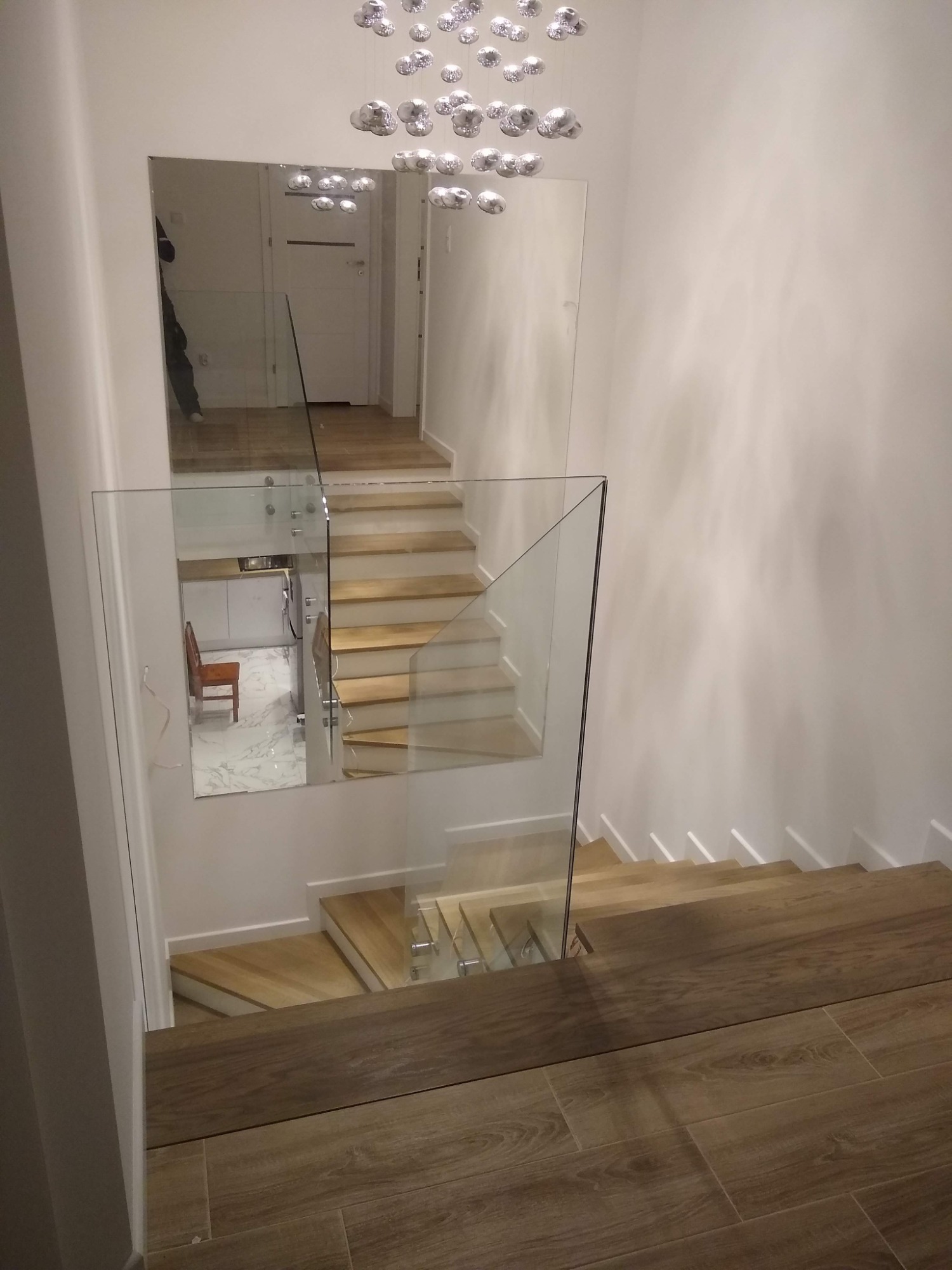 Szklana balustrada schodowa z widokiem na odbicie kuchni w lustrze, drewniane schody i podłoga z płytek imitujących drewno, nowoczesna lampa sufitowa.