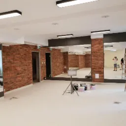 Wnętrze sali z ceglanymi ścianami, lustrami i rozstawionymi drabinami, przygotowywane do odświeżenia koloru podłogi, widoczne otwarte puszki z farbą oraz oświetlenie na suficie.