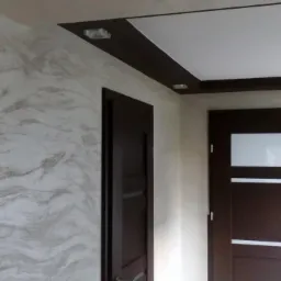 Wykończone wnętrze korytarza z dekoracyjną ścianą w odcieniach szarości i beżu, dwiema ciemnobrązowymi drzwiami z przeszkleniami, podwieszanym sufitem z oświetleniem punktowym i ciemną listwą.