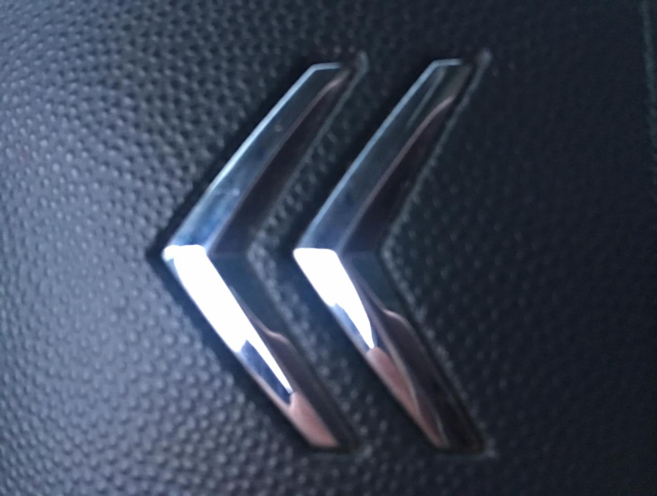 Błyszczące, chromowane logo Citroena na czarnej, teksturowanej powierzchni deski rozdzielczej pojazdu, z widocznymi refleksami światła.