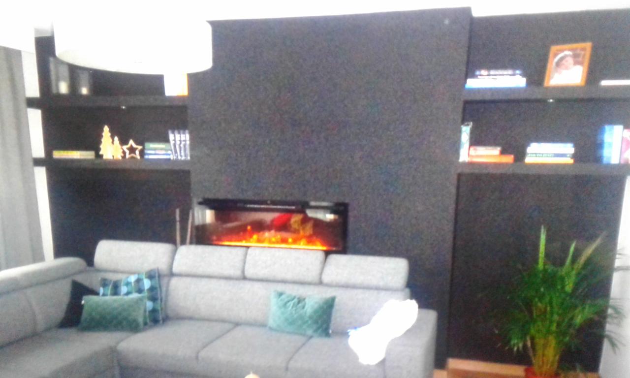 Szara sofa narożna z poduszkami w odcieniach zieleni i błękitu na tle czarnej ściany z wbudowanym elektrycznym kominkiem i półkami z dekoracjami.