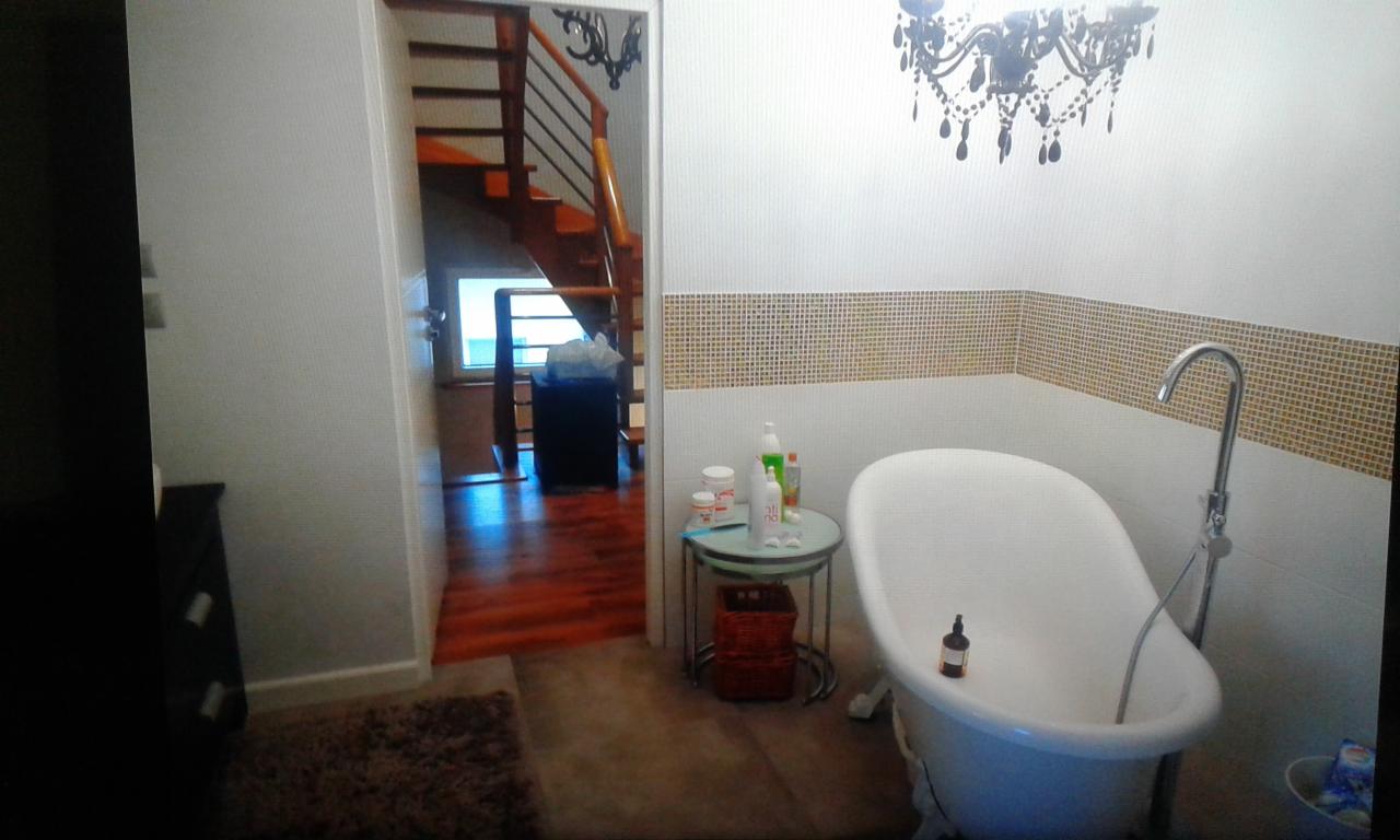 Biała wanna wolnostojąca z chromowaną armaturą na tle ściany z mozaiką, obok stolik z kosmetykami i widok na drewniane schody z balustradą.