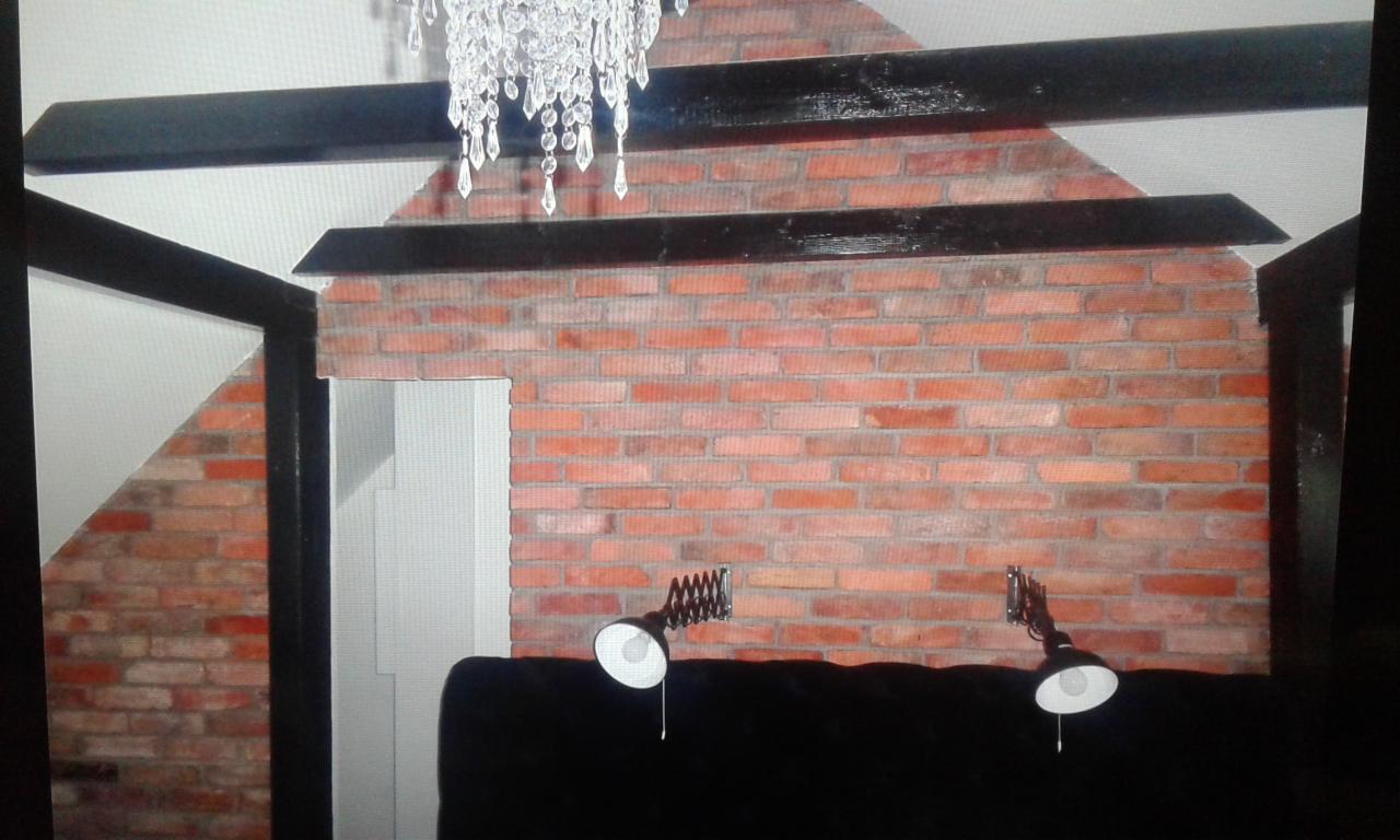 Fragment wnętrza z dekoracyjną ścianą z czerwonej cegły, czarnymi belkami stropowymi, kryształowym żyrandolem i dwoma lampkami ściennymi nad czarnym wezgłowiem łóżka.