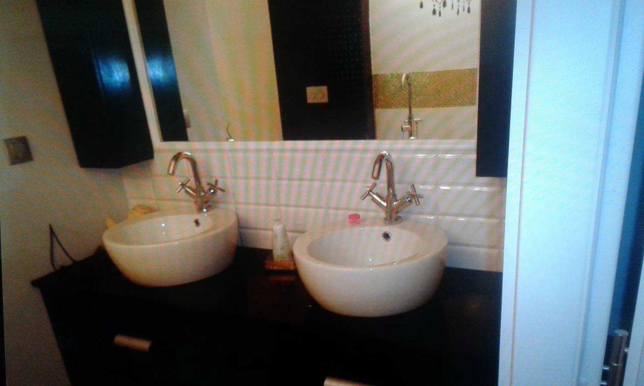 Dwie białe umywalki nablatowe z chromowanymi bateriami na czarnym blacie, białe płytki imitujące cegłę na ścianie, duże lustro z czarną szafką po lewej stronie i dekoracyjnym elementem nad płytkami.