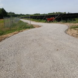 GRE-KOP Zygmunt Adam - Utwardzona, kręta droga szutrowa z pomarańczową taczką w tle, biegnąca wzdłuż ogrodzenia i paneli słonecznych. Widok na drzewa i niebo.