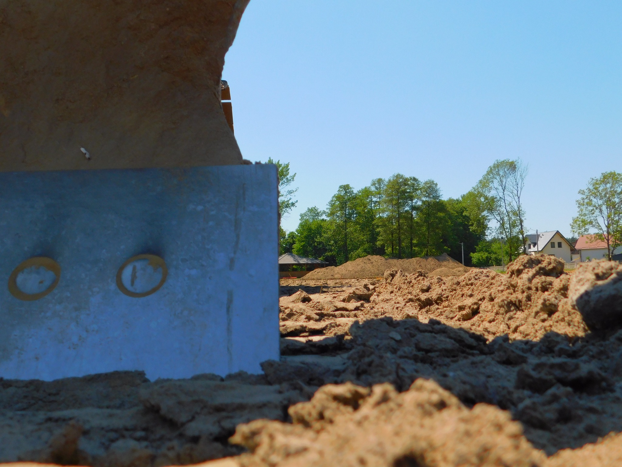 Niski kąt widzenia na hałdę ziemi z fragmentem metalowej płyty z otworami, zieleń drzew i zabudowania w tle, błękitne niebo. Teren po niwelacji.