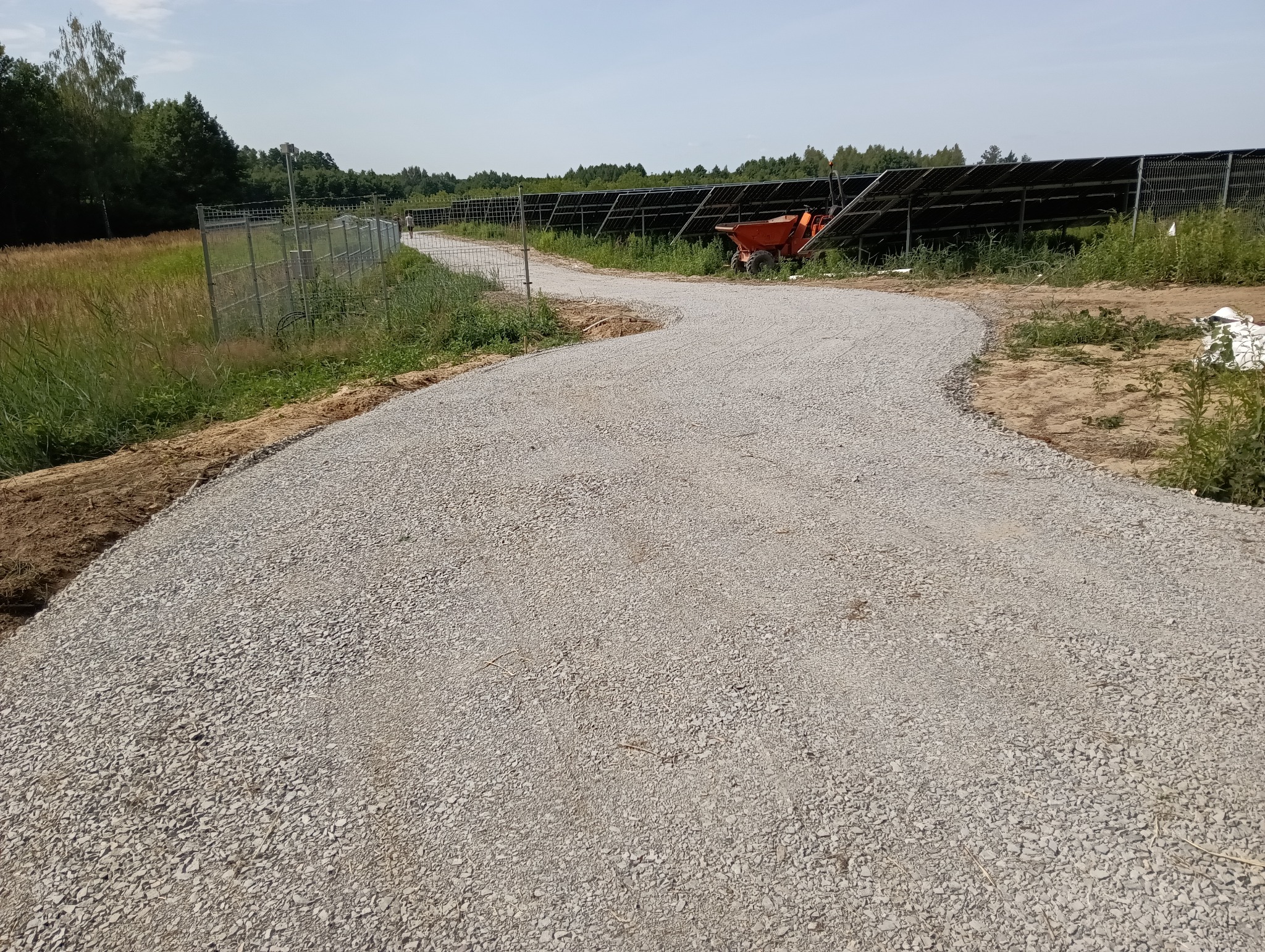 Utwardzona, kręta droga szutrowa z pomarańczową taczką w tle, biegnąca wzdłuż ogrodzenia i paneli słonecznych. Widok na drzewa i niebo.