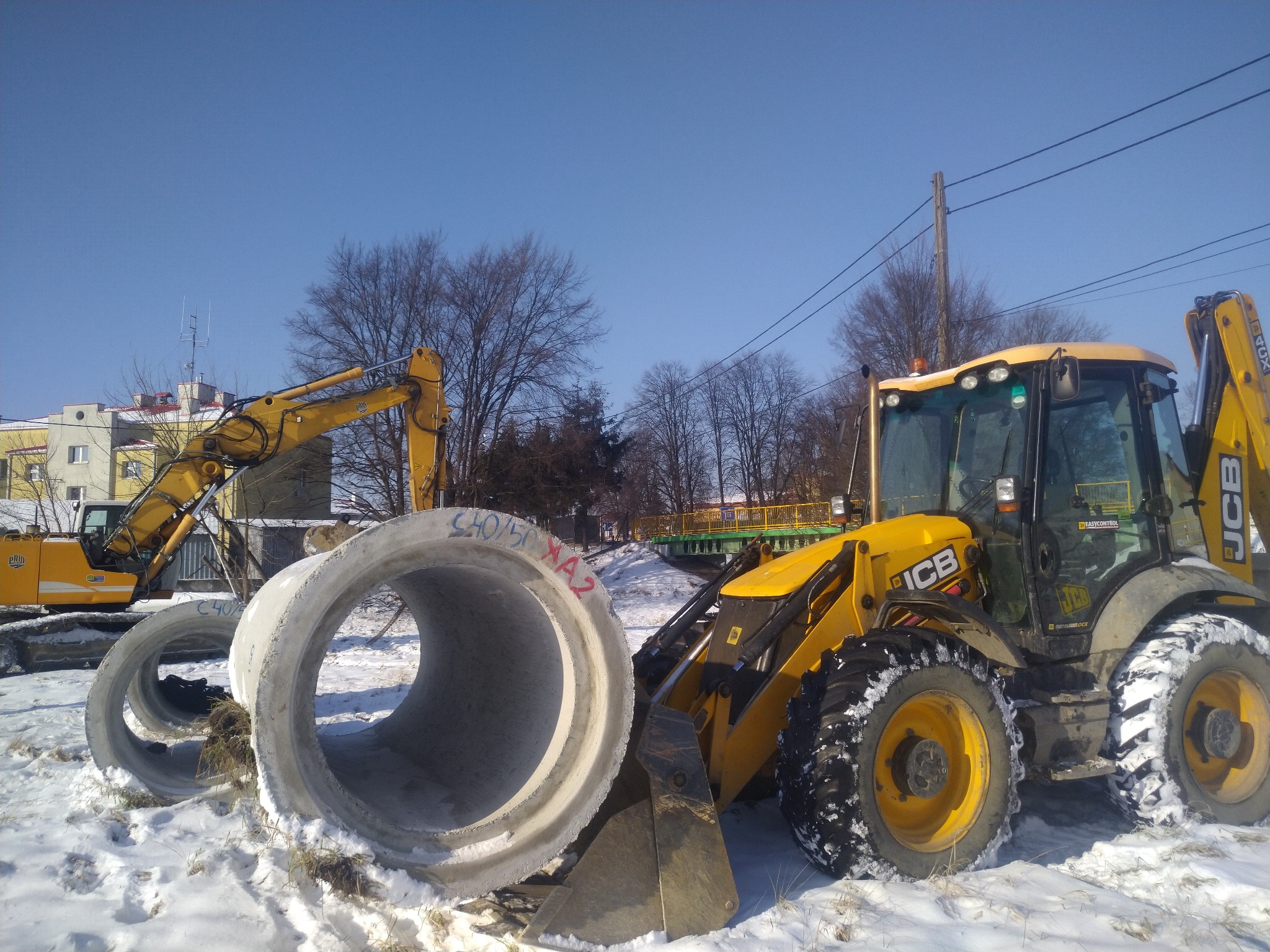 Żółta koparka JCB z betonowymi rurami na śniegu w Kolbuszowej, w tle druga koparka i budynek. Błękitne niebo.