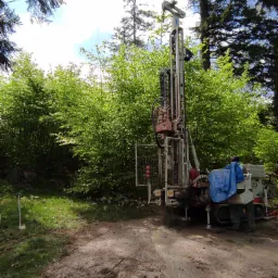 Wiertnica Comacchio GEO 305 w trakcie pracy na leśnej działce, widoczny operator w kaloszach i niebieskiej plandece, błotniste podłoże.