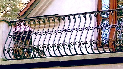 Kuta balustrada balkonowa z ozdobnymi elementami, widoczna z bliska, na tle jasnej elewacji budynku z oknem.