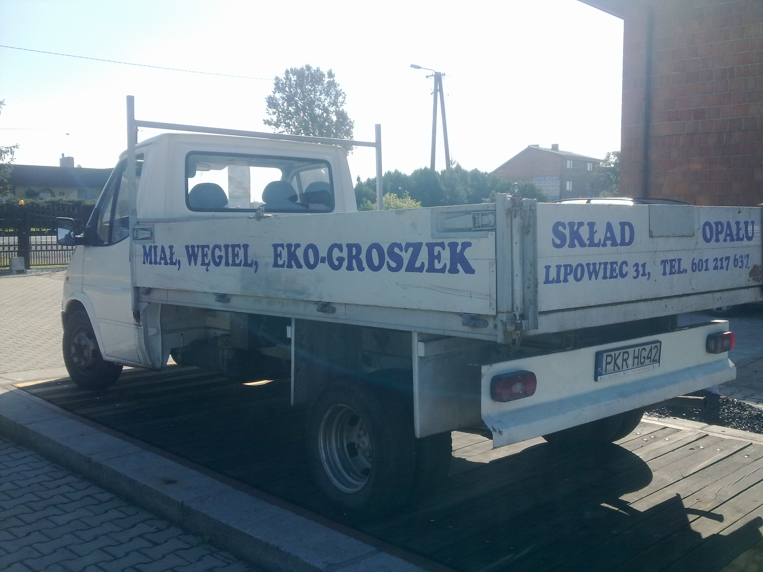 Biała dostawcza ciężarówka z niebieskim napisem 'Miał, Węgiel, Ekogroszek' i 'Skład Opału Lipowiec 31' na skrzyni ładunkowej, stojąca na wadze samochodowej.