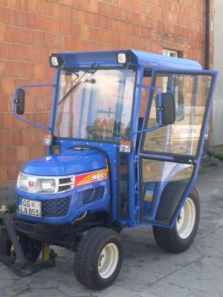 Niebieski ciągnik Iseki z otwartą kabiną z szybami, widoczna tablica rejestracyjna i ceglana ściana w tle.