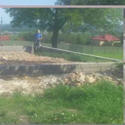 Budowa fundamentów domu jednorodzinnego z bloczków betonowych na działce z widokiem na okoliczne domy i drzewa, w tle zaparkowany bus.