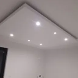 Biały sufit podwieszany z sześcioma punktowymi lampami LED, widoczne narożne wykończenie i fragment ściany, drabina w rogu pomieszczenia.