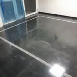 Błyszcząca, ciemnoszara posadzka z wyraźnie zaznaczonymi białymi fugami obwodowymi w nowo wykończonym pomieszczeniu. Widoczne odbicia światła na powierzchni płytek.