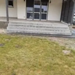 Nowoczesny, surowy taras z betonu architektonicznego przed domem jednorodzinnym w Grudziądzu. Widoczne schody prowadzące do drzwi balkonowych, trawnik z rzadką trawą i fragment elewacji budynku.