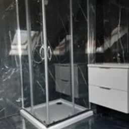 Kabina prysznicowa narożna z chromowanymi uchwytami na tle ściany z czarnego marmuru i białej szafki z umywalką.